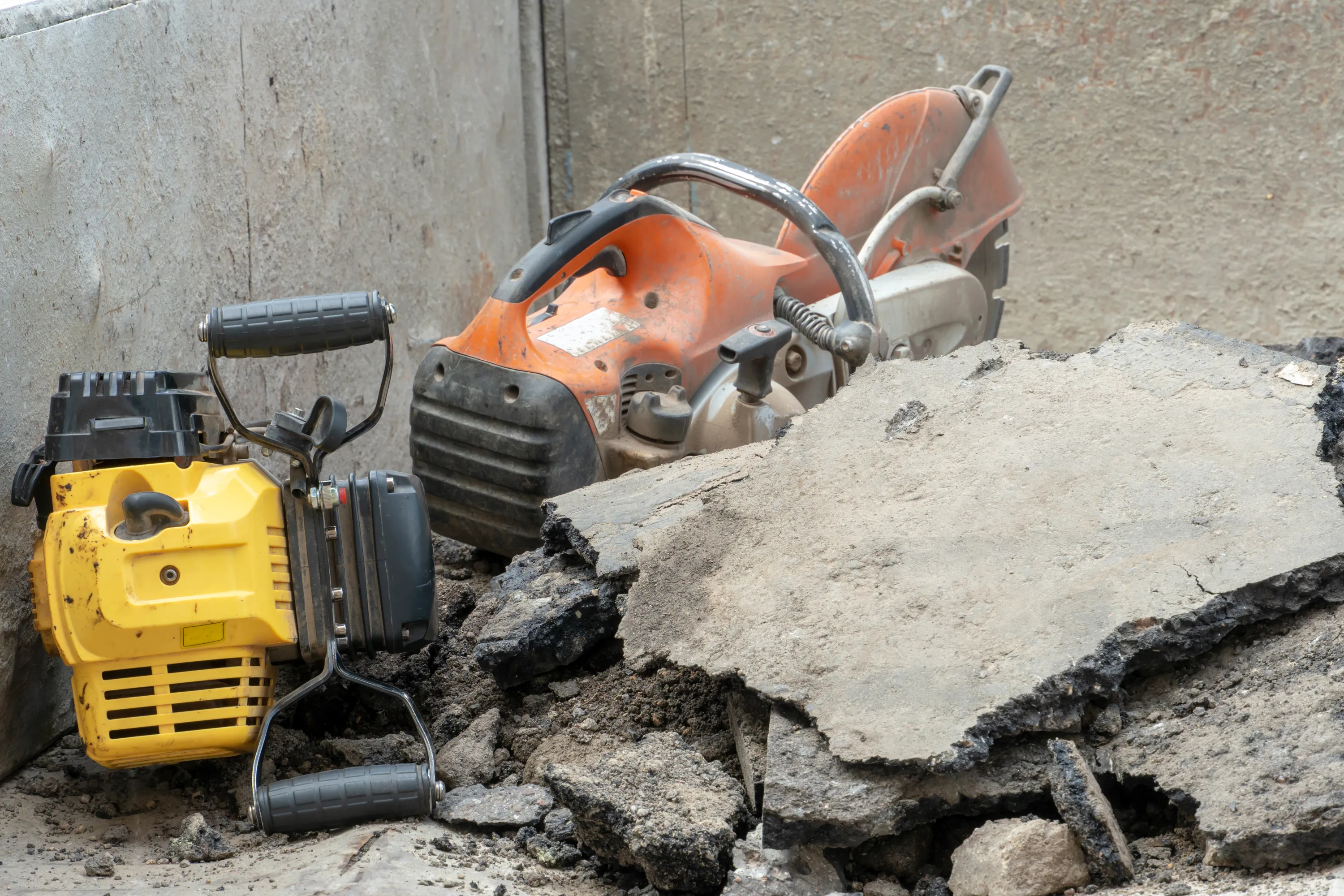 Keltainen piikkauskone ja oranssi katkaisusaha makaavat rikkinäisen betonin ja asfaltin kappaleiden keskellä. Taustalla näkyy harmaa betoniseinä.